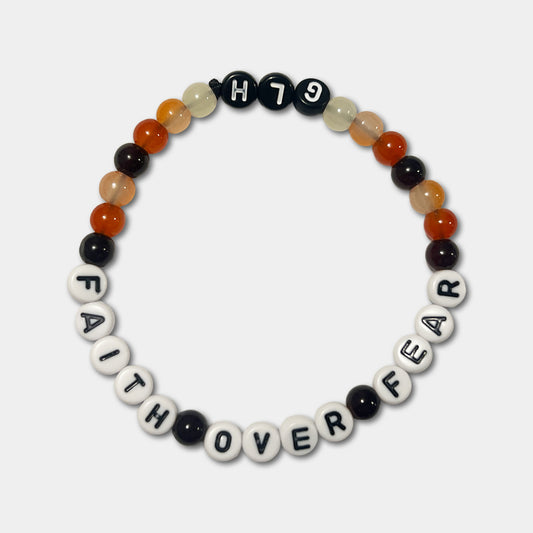 A beaded stretch bracelet arranged in a circle on a light gray background. The bracelet features white letter beads spelling 'FAITH OVER FEAR' with black lettering and an assortment of brown and orange beads.